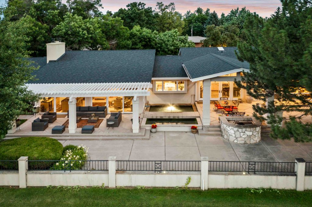 Outdoor living space of a luxury real estate home featuring a spacious covered patio with modern outdoor seating, a peaceful water feature, and a built-in stone grill, surrounded by lush greenery, perfect for entertaining or relaxing in style.