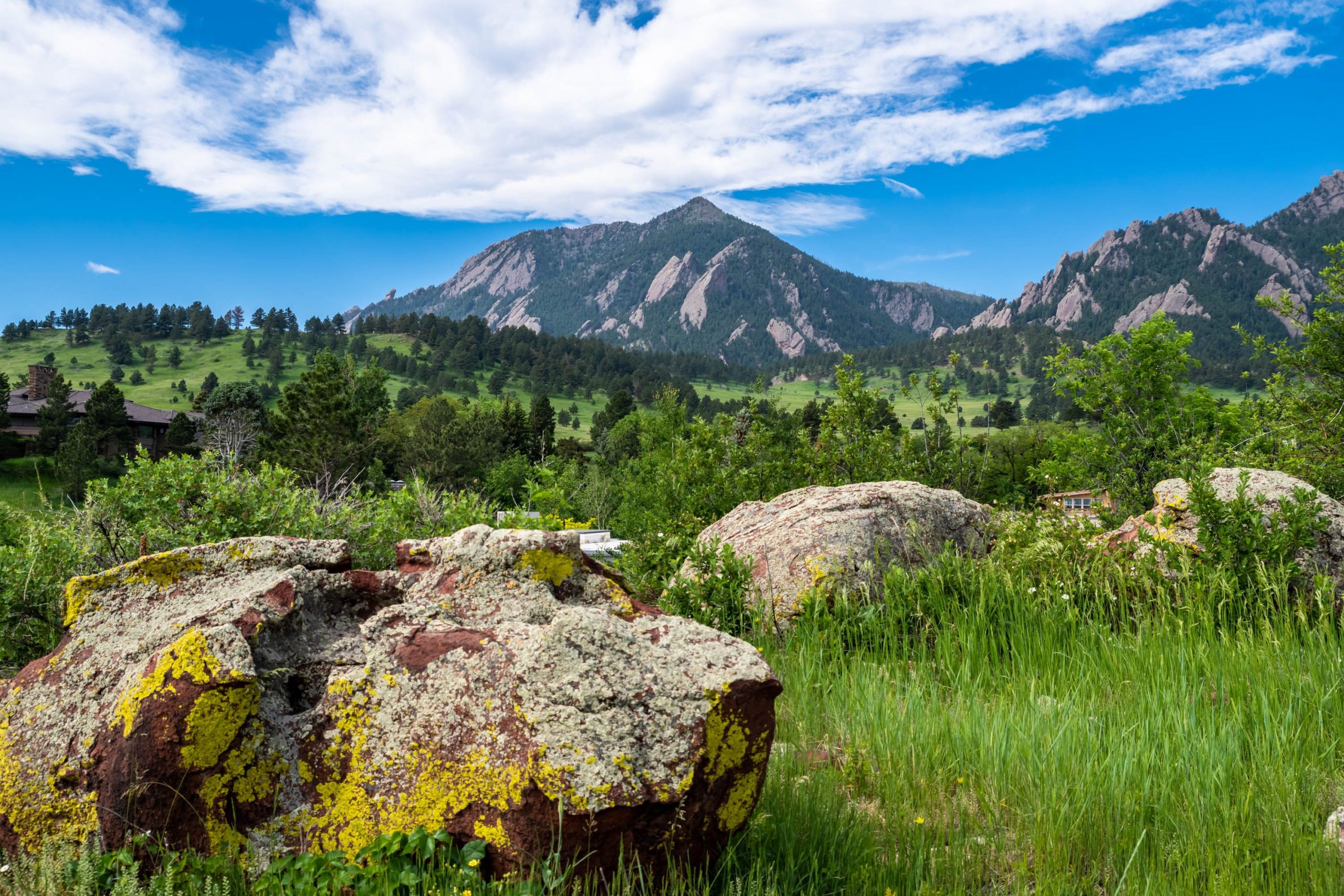 A scenic view of the Flatirons in Boulder, Colorado, with lush green hills, trees, and large boulders covered in yellow lichen in the foreground under a bright blue sky with scattered clouds.