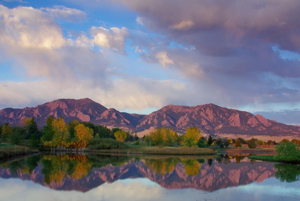 Scenic view of the Flatirons in Boulder, Colorado, reflecting in a calm lake surrounded by trees with autumn foliage under a partly cloudy sky.