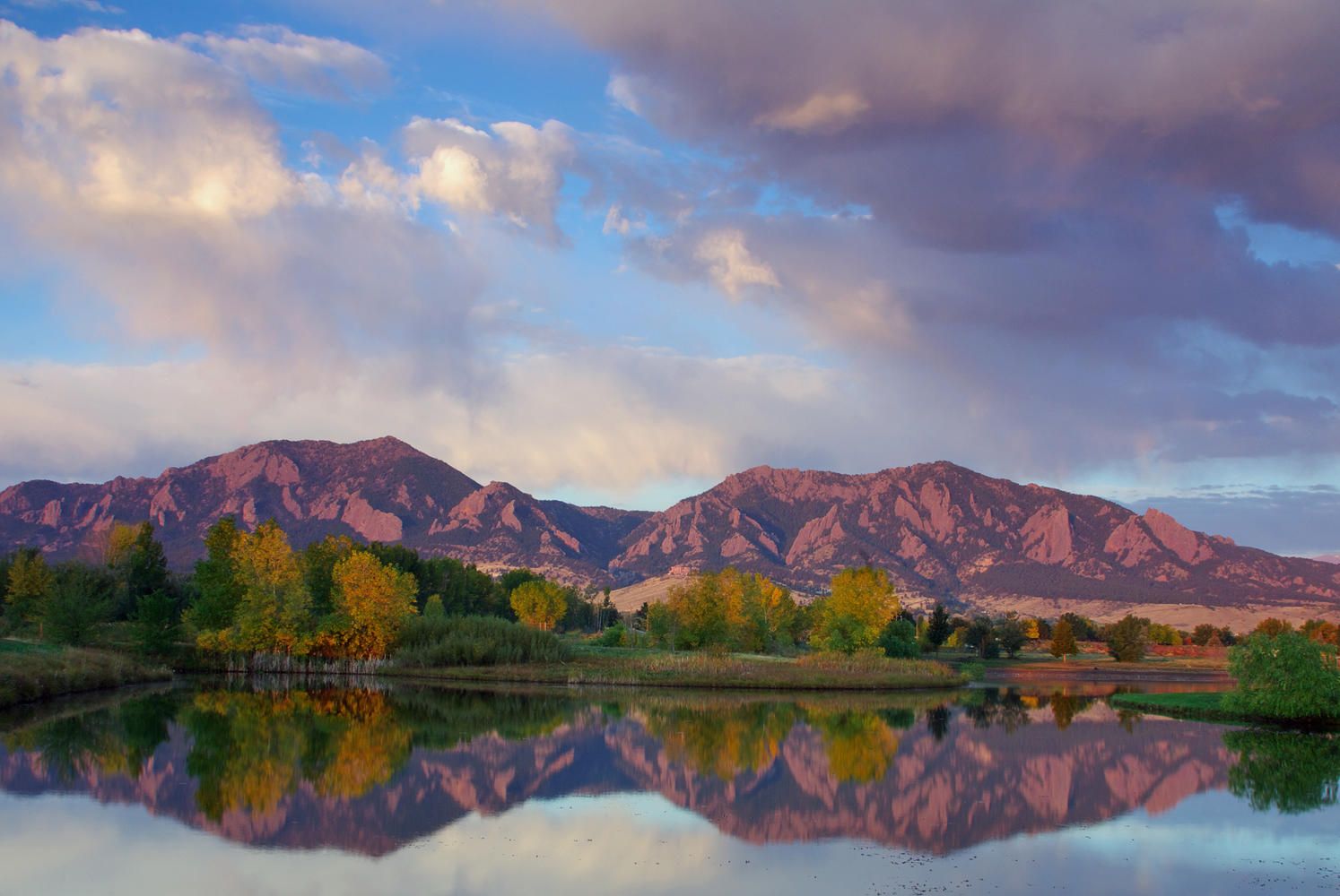 Scenic view of the Flatirons in Boulder, Colorado, reflecting in a calm lake surrounded by trees with autumn foliage under a partly cloudy sky.
