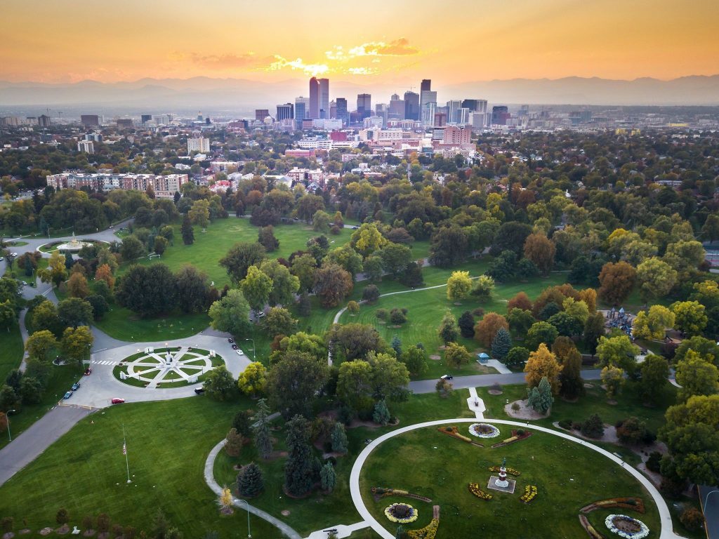 Aerial view of Denver, Colorado, at sunset with the city skyline in the background and a large park with green lawns and trees in the foreground.
