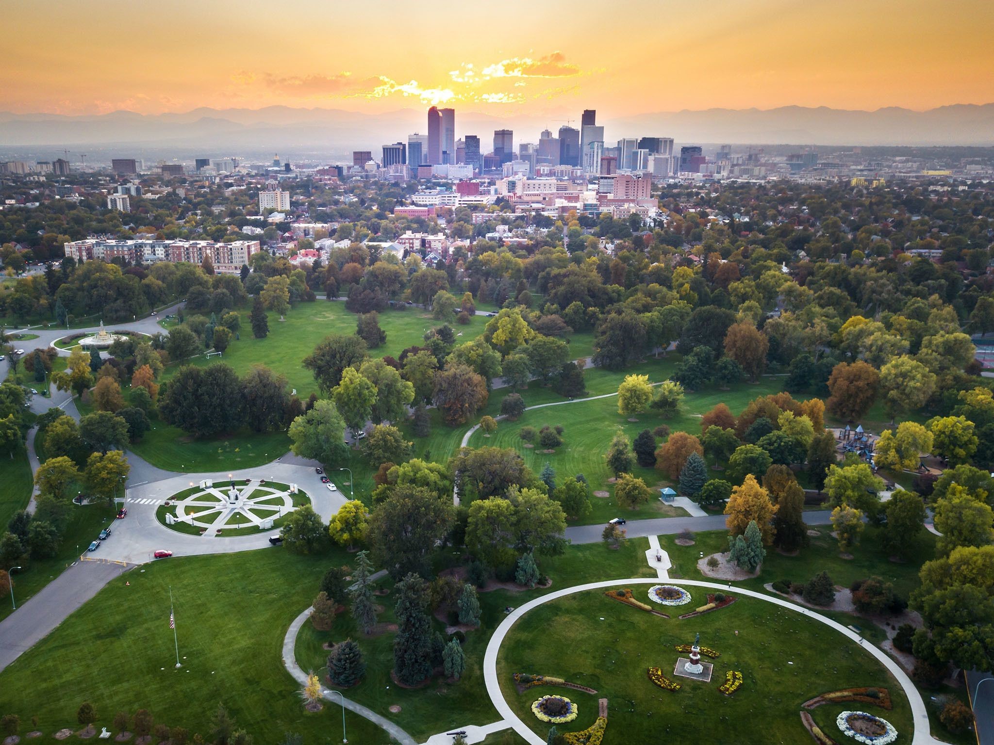 Aerial view of Denver, Colorado, at sunset with the city skyline in the background and a large park with green lawns and trees in the foreground.
